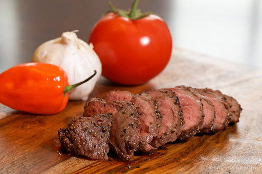 Bison sirloin on a cutting board with a tomato, habanero, and garlic bulb. Buy meat online from Apache.