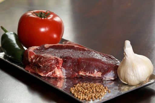 Organic bison ribeye from an online meat company. It rests on a serving tray with peppercorns, a garlic bulb, tomato, and jalapeno.